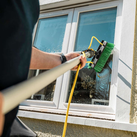 Person cleaning a glass window with a telescopic brush and water hose outside a building.